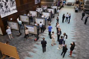 Image from upper level looking down on Accelerate to Industry event. Some participants stand beside poster boards while others chat together in small groups.