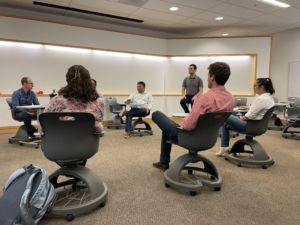 Students sitting in chairs within a classroom are focused on the panel of judges sitting at the front of the room. 