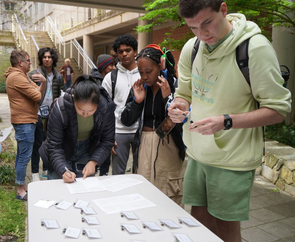 A group of students congregate around the name tag table, looking for their name tags.  One female student is writing her name on a sticky tag. In the background are professors and students chatting.