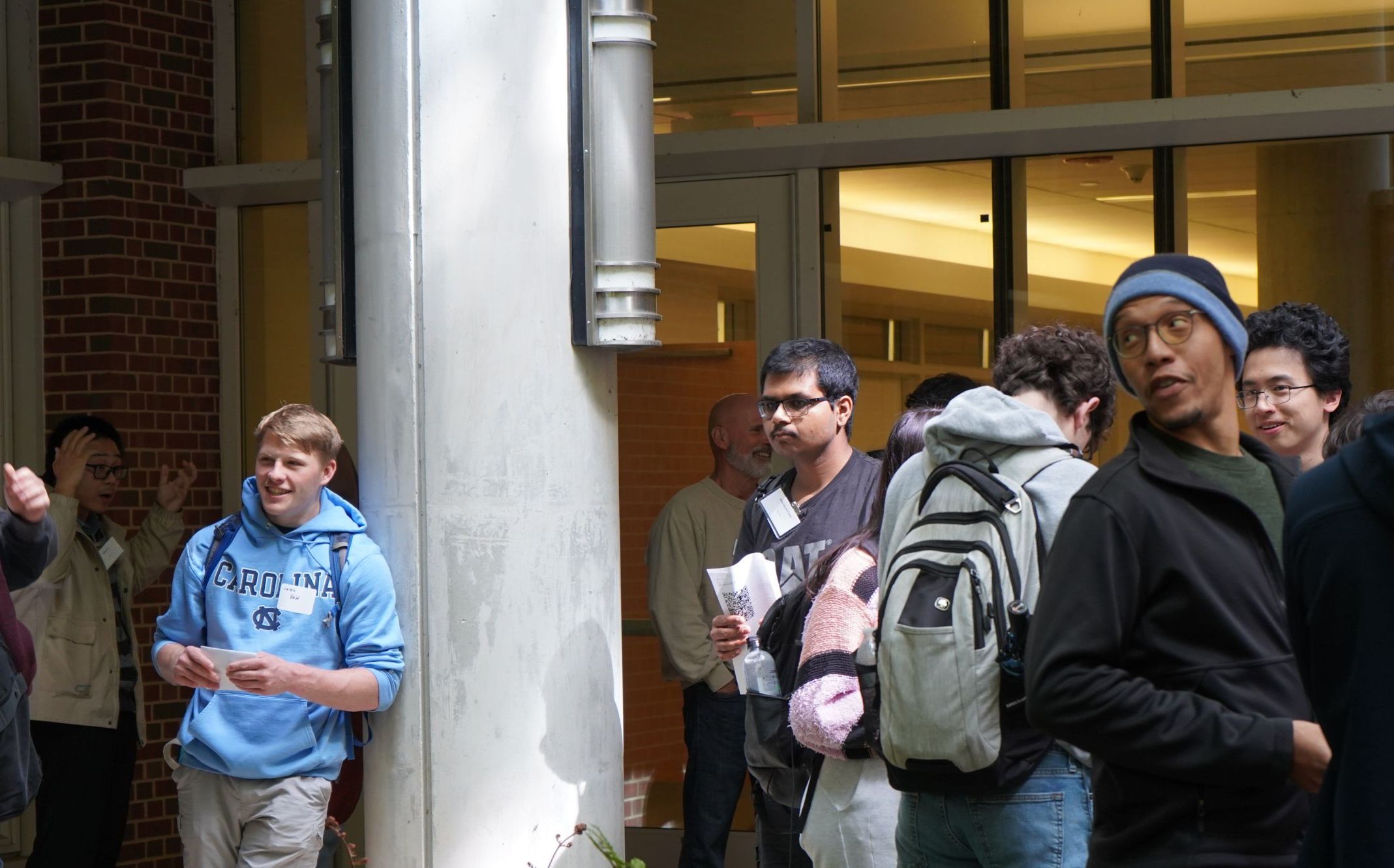 Professor Wilkerson-Hill turns towards something that has caught his attention behind him.  Other students and faculty can be seen talking in the background. One student leans against a support post while smiling.