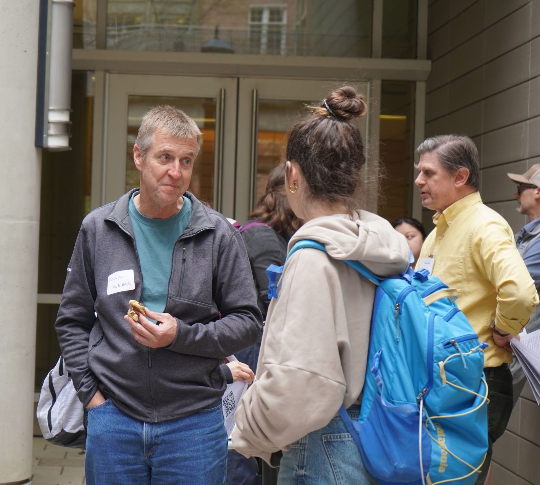 Professor Weeks smiles at a student while they talk about research. He is holding two cookies in his hand. Professor Tiani and Professor Hogan can be seen talking to students in the background.