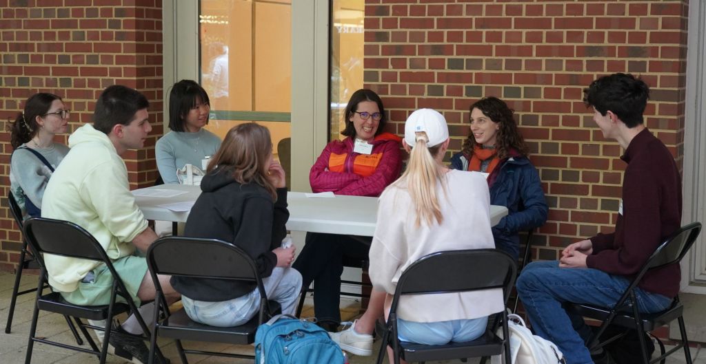 Professor Dempsey and Professor Jackson sit at a table with a group of students. Professor Jackson is in the middle of speaking to the students, while Professor Dempsey has possibly noticed the camera and is smiling in the direction of the photographer.