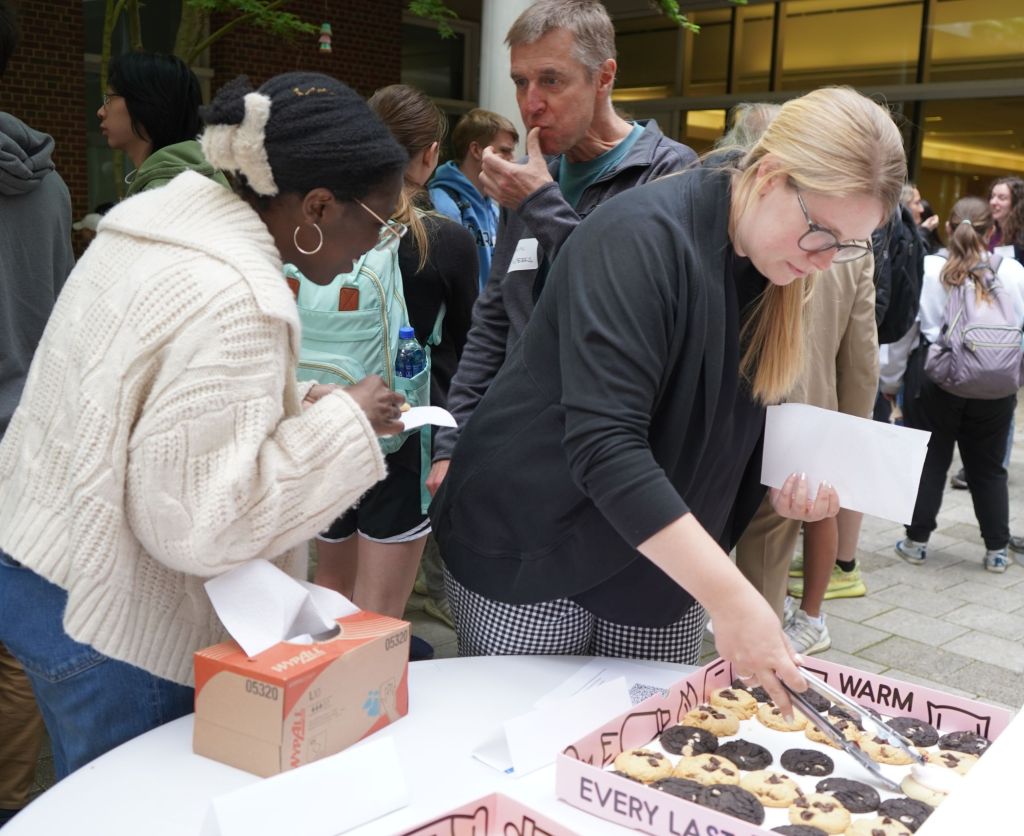 Two students select cookies from a tray while Professor Weeks walks behind them while licking his thumb. More students can be seen in the background.