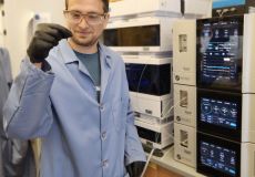Photo of Dr. Alex Zhukhovitskiy in a lab coat in front of some equipment. He is turning towards the camera while looking at a sample pinched between the pointer finger and thumb of his right hand.