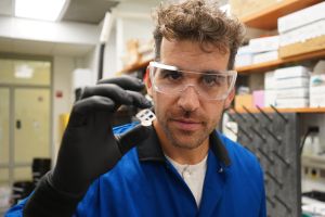 Libfarth, wearing a lab coat and goggles, showing to the camera a piece of polymer he collaborated with other researchers to create.